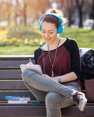 Eine junge Erwachsene sitzt an einem sonnigen Tag auf einer Bank im Park, hört Musik und schreibt Notizen.
